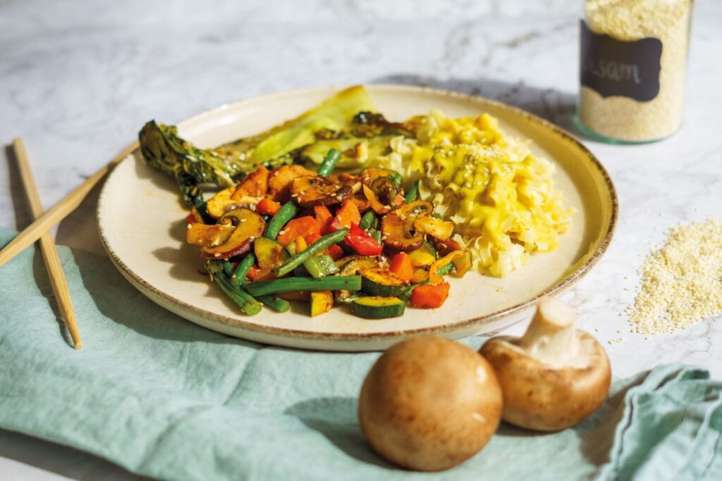 A vibrant vegetarian dish featuring grilled vegetables and mushrooms served with pasta, garnished with sesame seeds.