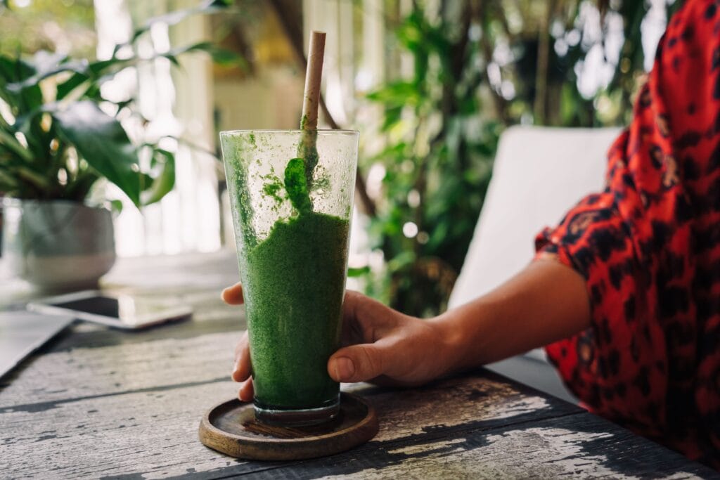 Hand holding a green smoothie in a glass on a rustic wooden table, surrounded by indoor plants.