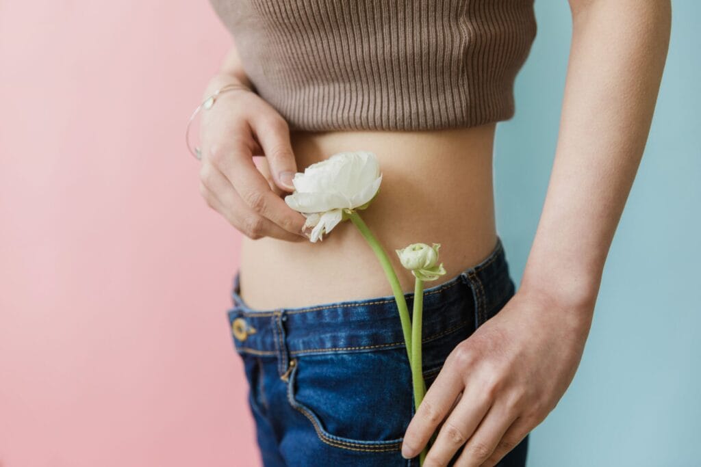 Fashionable woman in jeans and crop top holding a white flower against pink and blue backgrounds.