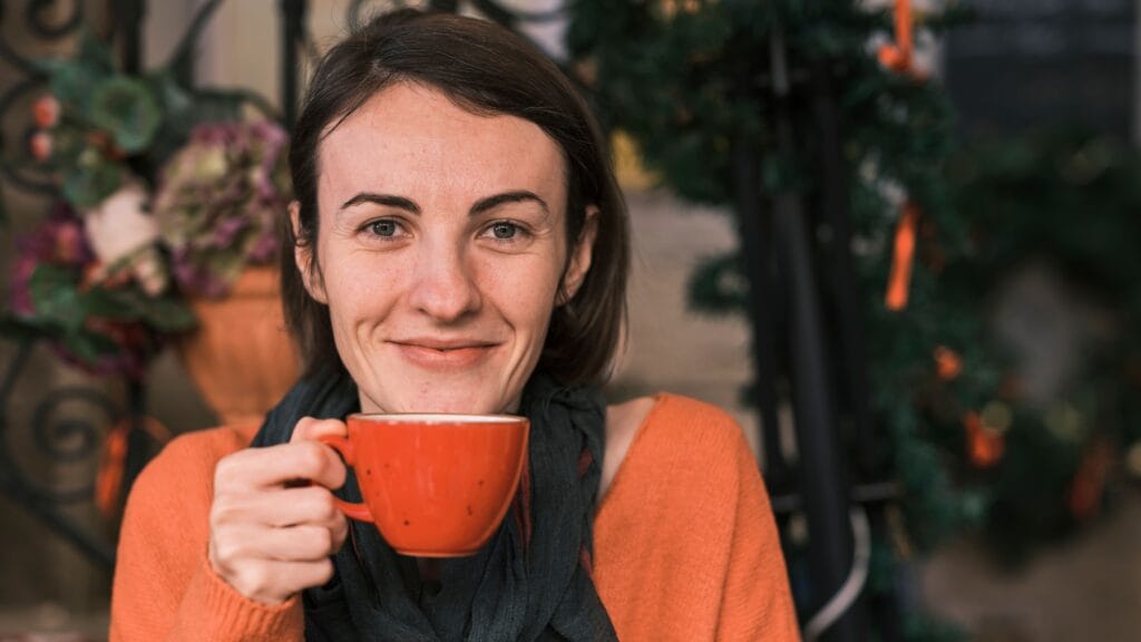 A cheerful woman in an orange sweater holds a warm drink, smiling while sitting outdoors.