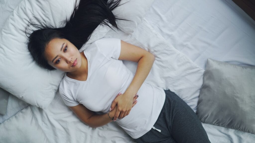 A young woman in casual attire lies on a white bed, looking relaxed and thoughtful.