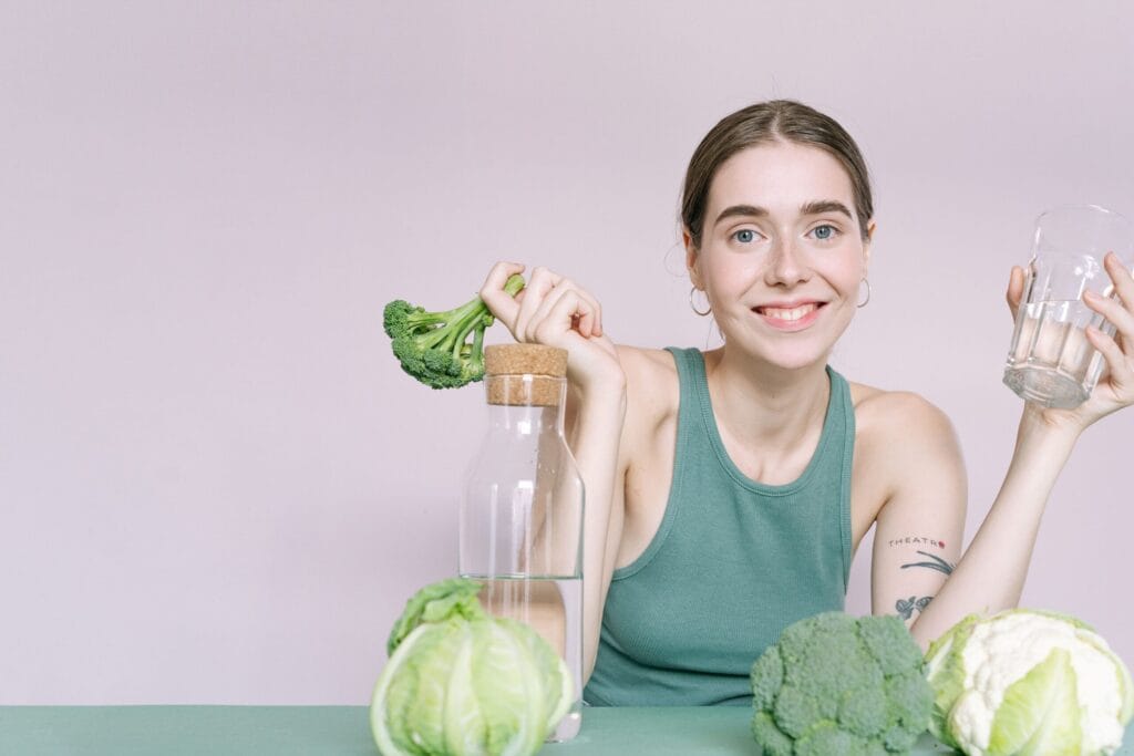 Woman in tank top smiling with broccoli, cabbage, and glass of water on table.