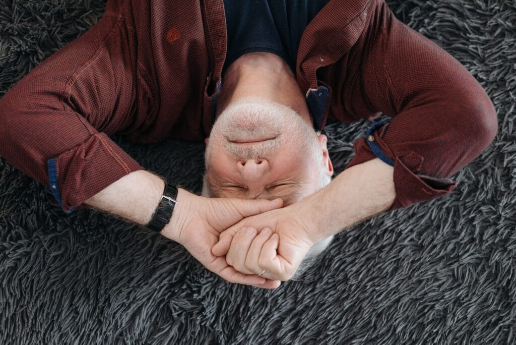A senior man lying on a soft surface indoors with closed eyes, expressing a range of emotions.
